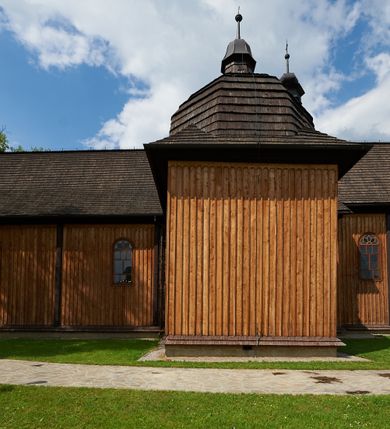 Zdjęcie nr 9: Kościół drewniany o konstrukcji zrębowej, na betonowym podmurowaniu przesłoniętym daszkiem gontowym. Elewacje jednolite, szalowane z listwowaniem, wzmocnione lisicami. Więźba dachowa drewniana, krokwiowa, o systemie więźbowo-zaskrzynieniowym. Kościół orientowany, jednonawowy, nawa na rzucie wydłużonego prostokąta, prezbiterium nieco węższe, zakończone trójbocznie. Do północnej ściany prezbiterium i nawy przylega zakrystia połączona z aneksem na rzucie prostokąta, do nawy od południa przylega duża kaplica na planie prostokąta otwarta do nawy otworem o wykroju prostokąta zamkniętego łukiem odcinkowym. 
Ściany wnętrza podzielone trzema parami pilastrów. Pomiędzy nawą a prezbiterium arkadowy otwór tęczowy z prostą belką. Parapet chóru muzycznego w zachodniej części nawy, oparty na dwóch kolumnach o wielobocznych trzonach. Wnętrze nakryte stropem płaskim, w nawie z zaskrzynieniami podtrzymywanymi przez trzy pary słupów o wielobocznych i kręconych trzonach. Otwory drzwiowe o prostokątnym wykroju zamknięte drzwiami jednoskrzydłowymi w północnej ścianie nawy oraz prezbiterium. Posadzka z płyt kamiennych. 
Elewacja frontowa jednoosiowa, zamknięta trójkątnym szczytem rozszerzającym się ku dołowi. Na osi ganek oparty na czterech słupach, nakryty dwuspadowym daszkiem gontowym, wejście do kościoła z dwuskrzydłowymi drzwiami. W partii szczytu dwa okna w formie stojącego prostokąta zamkniętego łukiem półkolistym. W elewacji południowej cztery otwory okienne w formie stojącego prostokąta zamkniętego łukiem półkolistym, dwa w nawie i dwa w prezbiterium oraz dwa analogiczne okna na zachodniej i wschodniej ścianie kaplicy. Elewacja północna z dwoma otworami okiennymi w formie stojącego prostokąta zamkniętego łukiem półkolistym, trzema oknami i wejściem do zakrystii i aneksu. Kościół nakryty wspólnym dachem o jednej kalenicy nad prezbiterium i nawą, gontowym, dwuspadowym, z przedłużoną połacią przekrywającą zaskrzynienia. W jednej trzeciej długości dachu nad nawą od strony prezbiterium wznosi się ponad kalenicą wieżyczka na sygnaturkę, na ośmiobocznym obitym blachą trzonie, z latarnią zwieńczoną niewielkim cebulastym hełmem. Kaplica boczna nakryta kopułą krytą gontem, z latarnią. Nad zakrystią i aneksem dach jednospadowy.
 
