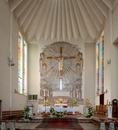 Zdjęcie nr 6: Świątynia trójnawowa, ośmioprzęsłowa o konstrukcji żelbetowej, tynkowana. Prezbiterium zamknięte prostą ścianą, umieszczone na kilkustopniowym podwyższeniu. Prostokątna zakrystia od strony wschodniej oraz prostokątna kaplica od strony zachodniej. Nawy, z których środkowa wyższa i szersza od bocznych wydzielone smukłymi filarami na rzucie prostokąta. Nakryte ekspresyjnym, żelbetowym stropem o rzeźbiarskiej formie. Chór muzyczny od strony południowej o konstrukcji żelbetowej z prostym parapetem zamkniętym od góry drewnianą poręczą. Fasada dwukondygnacyjna z umieszczonym w przyziemiu pięcioosiowym wejściem w formie prostokątnych otworów drzwiowych dostępnych wysokimi schodami. Otwory drzwiowe wydzielone żelbetowymi lizenami, podziały kontynuowane także w drugiej kondygnacji, gdzie w miejsce otworów drzwiowych wysokie, prostokątne okna. Elewacja zamknięta ścianą szczytową w kształcie trójkąta o nierównych bokach i zwieńczona prostym krzyżem. Od zachodu do elewacji dostawiona wieża dzwonna na rzucie trójkąta zwężającego się ku górze i nakryta strzelistym, ostrosłupowym hełmem zwieńczonym krzyżem. Elewacje boczne ośmioosiowe, artykułowane lizenami. W czterech pierwszych osiach od południa i na szerokości prezbiterium po dwa wysokie i prostokątne okna, w pozostałych pojedyncze okna i trójkątne szczyty. Od wschodu dach kryty blachą, dwuspadowy o nierównych połaciach; nad nawą boczną od strony wschodniej jednopołaciowy. Niewielka wieżyczka na sygnaturkę na kalenicy dachu od strony północnej.