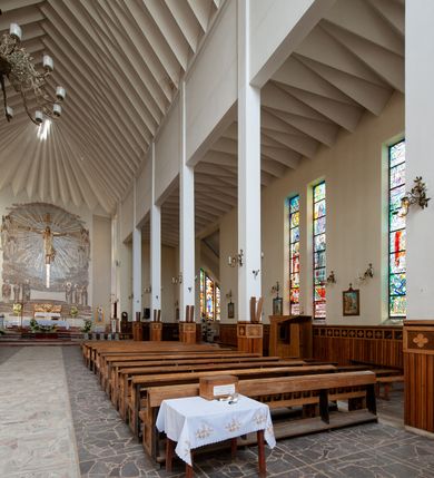 Zdjęcie nr 7: Świątynia trójnawowa, ośmioprzęsłowa o konstrukcji żelbetowej, tynkowana. Prezbiterium zamknięte prostą ścianą, umieszczone na kilkustopniowym podwyższeniu. Prostokątna zakrystia od strony wschodniej oraz prostokątna kaplica od strony zachodniej. Nawy, z których środkowa wyższa i szersza od bocznych wydzielone smukłymi filarami na rzucie prostokąta. Nakryte ekspresyjnym, żelbetowym stropem o rzeźbiarskiej formie. Chór muzyczny od strony południowej o konstrukcji żelbetowej z prostym parapetem zamkniętym od góry drewnianą poręczą. Fasada dwukondygnacyjna z umieszczonym w przyziemiu pięcioosiowym wejściem w formie prostokątnych otworów drzwiowych dostępnych wysokimi schodami. Otwory drzwiowe wydzielone żelbetowymi lizenami, podziały kontynuowane także w drugiej kondygnacji, gdzie w miejsce otworów drzwiowych wysokie, prostokątne okna. Elewacja zamknięta ścianą szczytową w kształcie trójkąta o nierównych bokach i zwieńczona prostym krzyżem. Od zachodu do elewacji dostawiona wieża dzwonna na rzucie trójkąta zwężającego się ku górze i nakryta strzelistym, ostrosłupowym hełmem zwieńczonym krzyżem. Elewacje boczne ośmioosiowe, artykułowane lizenami. W czterech pierwszych osiach od południa i na szerokości prezbiterium po dwa wysokie i prostokątne okna, w pozostałych pojedyncze okna i trójkątne szczyty. Od wschodu dach kryty blachą, dwuspadowy o nierównych połaciach; nad nawą boczną od strony wschodniej jednopołaciowy. Niewielka wieżyczka na sygnaturkę na kalenicy dachu od strony północnej.