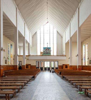 Zdjęcie nr 8: Świątynia trójnawowa, ośmioprzęsłowa o konstrukcji żelbetowej, tynkowana. Prezbiterium zamknięte prostą ścianą, umieszczone na kilkustopniowym podwyższeniu. Prostokątna zakrystia od strony wschodniej oraz prostokątna kaplica od strony zachodniej. Nawy, z których środkowa wyższa i szersza od bocznych wydzielone smukłymi filarami na rzucie prostokąta. Nakryte ekspresyjnym, żelbetowym stropem o rzeźbiarskiej formie. Chór muzyczny od strony południowej o konstrukcji żelbetowej z prostym parapetem zamkniętym od góry drewnianą poręczą. Fasada dwukondygnacyjna z umieszczonym w przyziemiu pięcioosiowym wejściem w formie prostokątnych otworów drzwiowych dostępnych wysokimi schodami. Otwory drzwiowe wydzielone żelbetowymi lizenami, podziały kontynuowane także w drugiej kondygnacji, gdzie w miejsce otworów drzwiowych wysokie, prostokątne okna. Elewacja zamknięta ścianą szczytową w kształcie trójkąta o nierównych bokach i zwieńczona prostym krzyżem. Od zachodu do elewacji dostawiona wieża dzwonna na rzucie trójkąta zwężającego się ku górze i nakryta strzelistym, ostrosłupowym hełmem zwieńczonym krzyżem. Elewacje boczne ośmioosiowe, artykułowane lizenami. W czterech pierwszych osiach od południa i na szerokości prezbiterium po dwa wysokie i prostokątne okna, w pozostałych pojedyncze okna i trójkątne szczyty. Od wschodu dach kryty blachą, dwuspadowy o nierównych połaciach; nad nawą boczną od strony wschodniej jednopołaciowy. Niewielka wieżyczka na sygnaturkę na kalenicy dachu od strony północnej.