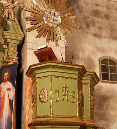 Zdjęcie nr 1: Ambona przyścienna, wisząca. Kosz na planie prostokąta o ściętych narożach, z dobudowanym po prawej stronie balkonem, opasującym ścianę tęczową, prowadzącym do drzwi umieszczonych w północnej ścianie prezbiterium. Kosz ze schodkowym i profilowanym podwieszeniem zakończonym szyszką. Kosz i balkon na profilowanych cokołach, o ściankach dekorowanych płycinami z perełkowaniem, w polu frontowym monogram IHS, w czterech polach flankujących rozety. Ścianki zakończone profilowanym gzymsem stanowiącym parapet. Ponad koszem, na podłuczu arkady tęczowej zawieszona  gloria promienista z gołębicą Ducha Świętego w obłokach.
Struktura polichromowana w kolorze jasnozielonym, dekoracja, profile i gloria złocone, gołębica i obłoki srebrzone.


