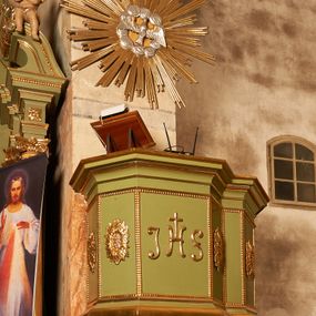 Zdjęcie nr 1: Ambona przyścienna, wisząca. Kosz na planie prostokąta o ściętych narożach, z dobudowanym po prawej stronie balkonem, opasującym ścianę tęczową, prowadzącym do drzwi umieszczonych w północnej ścianie prezbiterium. Kosz ze schodkowym i profilowanym podwieszeniem zakończonym szyszką. Kosz i balkon na profilowanych cokołach, o ściankach dekorowanych płycinami z perełkowaniem, w polu frontowym monogram IHS, w czterech polach flankujących rozety. Ścianki zakończone profilowanym gzymsem stanowiącym parapet. Ponad koszem, na podłuczu arkady tęczowej zawieszona  gloria promienista z gołębicą Ducha Świętego w obłokach.
Struktura polichromowana w kolorze jasnozielonym, dekoracja, profile i gloria złocone, gołębica i obłoki srebrzone.


