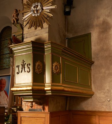 Zdjęcie nr 2: Ambona przyścienna, wisząca. Kosz na planie prostokąta o ściętych narożach, z dobudowanym po prawej stronie balkonem, opasującym ścianę tęczową, prowadzącym do drzwi umieszczonych w północnej ścianie prezbiterium. Kosz ze schodkowym i profilowanym podwieszeniem zakończonym szyszką. Kosz i balkon na profilowanych cokołach, o ściankach dekorowanych płycinami z perełkowaniem, w polu frontowym monogram IHS, w czterech polach flankujących rozety. Ścianki zakończone profilowanym gzymsem stanowiącym parapet. Ponad koszem, na podłuczu arkady tęczowej zawieszona  gloria promienista z gołębicą Ducha Świętego w obłokach.
Struktura polichromowana w kolorze jasnozielonym, dekoracja, profile i gloria złocone, gołębica i obłoki srebrzone.


