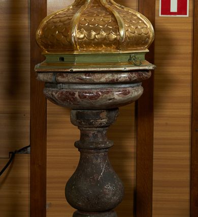 Zdjęcie nr 2: Chrzcielnica na masywnej kamiennej podstawie na planie ściętego koła. Trzon z piaskowca, polichromowany na sposób imitujący marmoryzację, w formie jednolalkowej tralki z okrągłą plintą i sześcienną bazą. Czara marmurowa w formie spłaszczonej półkuli, z wywiniętym brzegiem, ujęta metalowymi cęgami z kotwami. Wewnątrz blaszana misa. 
Pokrywa drewniana, na profilowanej podstawie na planie ośmioboku, w formie kopułki pokrytej łuską, podzielonej na osiem pól, zwieńczona cokołem z niewielką grupą rzeźbiarską Chrztu Chrystusa. Pokrywa i profile złocone, podstawa polichromowana w kolorze zielonym. 




