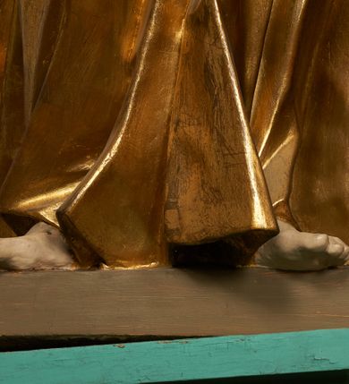 Zdjęcie nr 5: Rzeźba drążona z tyłu, ustawiona na niskiej podstawie. Figura zwrócona jest frontalnie, z głową skierowaną w lewo, w kontrapoście, o odchylonej i esowato wygiętej sylwetce. Twarz ma szczupłą, o migdałowatych, głęboko osadzonych oczach, prostym nosie, wąskich ustach, z uwypukloną brodą, okoloną jasnymi, falowanymi włosami ukrytymi pod welonem. Święta ubrana jest w przylegającą do ciała złoconą tunikę przepasaną w talii, na lewe ramię ma narzucony płaszcz, opadający po bokach postaci z fragmentem tkaniny zarzuconym na lewą nogę, na głowie ma welon, rozwiany na plecy. Ciało polichromowane naturalistycznie.
