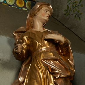 Zdjęcie nr 1: Rzeźba drążona z tyłu, ustawiona na niskiej podstawie. Figura zwrócona jest frontalnie, z głową skierowaną w lewo, w kontrapoście, o odchylonej i esowato wygiętej sylwetce. Twarz ma szczupłą, o migdałowatych, głęboko osadzonych oczach, prostym nosie, wąskich ustach, z uwypukloną brodą, okoloną jasnymi, falowanymi włosami ukrytymi pod welonem. Święta ubrana jest w przylegającą do ciała złoconą tunikę przepasaną w talii, na lewe ramię ma narzucony płaszcz, opadający po bokach postaci z fragmentem tkaniny zarzuconym na lewą nogę, na głowie ma welon, rozwiany na plecy. Ciało polichromowane naturalistycznie.
