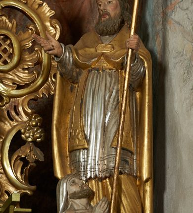 Zdjęcie nr 1: Rzeźba jest drążona i ścięta z tyłu, ustawiona na niskiej podstawie na planie kwadratu. Święty zwrócony jest frontalnie, z głową skierowaną w prawo, w kontrapoście, o odchylonej delikatnie sylwetce. Obie ręce ugięte, rozłożone na boki, w lewej trzyma pastorał, prawą unosi w geście błogosławieństwa.  Twarz ma pociągłą, szczupłą, o migdałowatych oczach, uniesionych brwiach, dużym nosie i pełnych, rozchylonych ustach, okoloną falowaną brodą. Włosy ma brązowe, falowane, sięgające ramion. Na głowie nosi srebrzoną i złoconą infułę z krzyżem kawalerskim w glorii. Święty ubrany jest w złoconą albę, srebrzoną rokietę i złoconą stułę, przepasane w talii. Na piersi ma pektorał, na ramionach zarzuconą złoconą kapę, zapiętą z przodu. Na stopach ma złocone buty. Przy prawej nodze świętego półpostać Piotrowina, zwróconego profilem, z uniesioną głową i dłońmi złożonymi w geście modlitwy. Twarz szczupła, z oczami skierowanymi w górę, okolona krótką brodą. Głowę ma nakrytą srebrzonym całunem opadającym na plecy, tors ma nagi. Polichromia ciała naturalistyczna.
