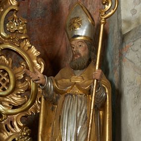 Zdjęcie nr 1: Rzeźba jest drążona i ścięta z tyłu, ustawiona na niskiej podstawie na planie kwadratu. Święty zwrócony jest frontalnie, z głową skierowaną w prawo, w kontrapoście, o odchylonej delikatnie sylwetce. Obie ręce ugięte, rozłożone na boki, w lewej trzyma pastorał, prawą unosi w geście błogosławieństwa.  Twarz ma pociągłą, szczupłą, o migdałowatych oczach, uniesionych brwiach, dużym nosie i pełnych, rozchylonych ustach, okoloną falowaną brodą. Włosy ma brązowe, falowane, sięgające ramion. Na głowie nosi srebrzoną i złoconą infułę z krzyżem kawalerskim w glorii. Święty ubrany jest w złoconą albę, srebrzoną rokietę i złoconą stułę, przepasane w talii. Na piersi ma pektorał, na ramionach zarzuconą złoconą kapę, zapiętą z przodu. Na stopach ma złocone buty. Przy prawej nodze świętego półpostać Piotrowina, zwróconego profilem, z uniesioną głową i dłońmi złożonymi w geście modlitwy. Twarz szczupła, z oczami skierowanymi w górę, okolona krótką brodą. Głowę ma nakrytą srebrzonym całunem opadającym na plecy, tors ma nagi. Polichromia ciała naturalistyczna.

