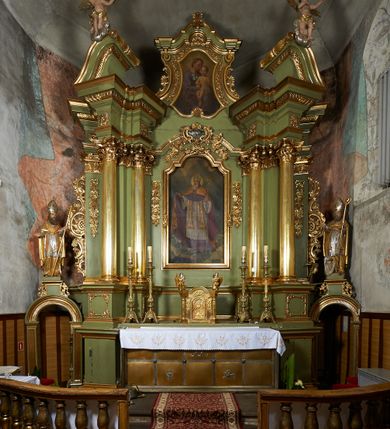 Zdjęcie nr 6: Rzeźba jest drążona i ścięta z tyłu, ustawiona na niskiej podstawie na planie kwadratu. Święty zwrócony jest frontalnie, z głową skierowaną w prawo, w kontrapoście, o odchylonej delikatnie sylwetce. Obie ręce ugięte, rozłożone na boki, w lewej trzyma pastorał, prawą unosi w geście błogosławieństwa.  Twarz ma pociągłą, szczupłą, o migdałowatych oczach, uniesionych brwiach, dużym nosie i pełnych, rozchylonych ustach, okoloną falowaną brodą. Włosy ma brązowe, falowane, sięgające ramion. Na głowie nosi srebrzoną i złoconą infułę z krzyżem kawalerskim w glorii. Święty ubrany jest w złoconą albę, srebrzoną rokietę i złoconą stułę, przepasane w talii. Na piersi ma pektorał, na ramionach zarzuconą złoconą kapę, zapiętą z przodu. Na stopach ma złocone buty. Przy prawej nodze świętego półpostać Piotrowina, zwróconego profilem, z uniesioną głową i dłońmi złożonymi w geście modlitwy. Twarz szczupła, z oczami skierowanymi w górę, okolona krótką brodą. Głowę ma nakrytą srebrzonym całunem opadającym na plecy, tors ma nagi. Polichromia ciała naturalistyczna.
