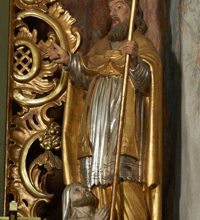 Zdjęcie nr 9: Rzeźba jest drążona i ścięta z tyłu, ustawiona na niskiej podstawie na planie kwadratu. Święty zwrócony jest frontalnie, z głową skierowaną w prawo, w kontrapoście, o odchylonej delikatnie sylwetce. Obie ręce ugięte, rozłożone na boki, w lewej trzyma pastorał, prawą unosi w geście błogosławieństwa.  Twarz ma pociągłą, szczupłą, o migdałowatych oczach, uniesionych brwiach, dużym nosie i pełnych, rozchylonych ustach, okoloną falowaną brodą. Włosy ma brązowe, falowane, sięgające ramion. Na głowie nosi srebrzoną i złoconą infułę z krzyżem kawalerskim w glorii. Święty ubrany jest w złoconą albę, srebrzoną rokietę i złoconą stułę, przepasane w talii. Na piersi ma pektorał, na ramionach zarzuconą złoconą kapę, zapiętą z przodu. Na stopach ma złocone buty. Przy prawej nodze świętego półpostać Piotrowina, zwróconego profilem, z uniesioną głową i dłońmi złożonymi w geście modlitwy. Twarz szczupła, z oczami skierowanymi w górę, okolona krótką brodą. Głowę ma nakrytą srebrzonym całunem opadającym na plecy, tors ma nagi. Polichromia ciała naturalistyczna.
