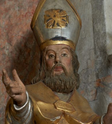 Zdjęcie nr 15: Rzeźba jest drążona i ścięta z tyłu, ustawiona na niskiej podstawie na planie kwadratu. Święty zwrócony jest frontalnie, z głową skierowaną w prawo, w kontrapoście, o odchylonej delikatnie sylwetce. Obie ręce ugięte, rozłożone na boki, w lewej trzyma pastorał, prawą unosi w geście błogosławieństwa.  Twarz ma pociągłą, szczupłą, o migdałowatych oczach, uniesionych brwiach, dużym nosie i pełnych, rozchylonych ustach, okoloną falowaną brodą. Włosy ma brązowe, falowane, sięgające ramion. Na głowie nosi srebrzoną i złoconą infułę z krzyżem kawalerskim w glorii. Święty ubrany jest w złoconą albę, srebrzoną rokietę i złoconą stułę, przepasane w talii. Na piersi ma pektorał, na ramionach zarzuconą złoconą kapę, zapiętą z przodu. Na stopach ma złocone buty. Przy prawej nodze świętego półpostać Piotrowina, zwróconego profilem, z uniesioną głową i dłońmi złożonymi w geście modlitwy. Twarz szczupła, z oczami skierowanymi w górę, okolona krótką brodą. Głowę ma nakrytą srebrzonym całunem opadającym na plecy, tors ma nagi. Polichromia ciała naturalistyczna.
