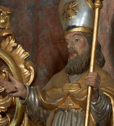 Zdjęcie nr 14: Rzeźba jest drążona i ścięta z tyłu, ustawiona na niskiej podstawie na planie kwadratu. Święty zwrócony jest frontalnie, z głową skierowaną w prawo, w kontrapoście, o odchylonej delikatnie sylwetce. Obie ręce ugięte, rozłożone na boki, w lewej trzyma pastorał, prawą unosi w geście błogosławieństwa.  Twarz ma pociągłą, szczupłą, o migdałowatych oczach, uniesionych brwiach, dużym nosie i pełnych, rozchylonych ustach, okoloną falowaną brodą. Włosy ma brązowe, falowane, sięgające ramion. Na głowie nosi srebrzoną i złoconą infułę z krzyżem kawalerskim w glorii. Święty ubrany jest w złoconą albę, srebrzoną rokietę i złoconą stułę, przepasane w talii. Na piersi ma pektorał, na ramionach zarzuconą złoconą kapę, zapiętą z przodu. Na stopach ma złocone buty. Przy prawej nodze świętego półpostać Piotrowina, zwróconego profilem, z uniesioną głową i dłońmi złożonymi w geście modlitwy. Twarz szczupła, z oczami skierowanymi w górę, okolona krótką brodą. Głowę ma nakrytą srebrzonym całunem opadającym na plecy, tors ma nagi. Polichromia ciała naturalistyczna.
