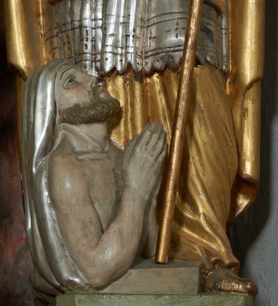 Zdjęcie nr 4: Rzeźba jest drążona i ścięta z tyłu, ustawiona na niskiej podstawie na planie kwadratu. Święty zwrócony jest frontalnie, z głową skierowaną w prawo, w kontrapoście, o odchylonej delikatnie sylwetce. Obie ręce ugięte, rozłożone na boki, w lewej trzyma pastorał, prawą unosi w geście błogosławieństwa.  Twarz ma pociągłą, szczupłą, o migdałowatych oczach, uniesionych brwiach, dużym nosie i pełnych, rozchylonych ustach, okoloną falowaną brodą. Włosy ma brązowe, falowane, sięgające ramion. Na głowie nosi srebrzoną i złoconą infułę z krzyżem kawalerskim w glorii. Święty ubrany jest w złoconą albę, srebrzoną rokietę i złoconą stułę, przepasane w talii. Na piersi ma pektorał, na ramionach zarzuconą złoconą kapę, zapiętą z przodu. Na stopach ma złocone buty. Przy prawej nodze świętego półpostać Piotrowina, zwróconego profilem, z uniesioną głową i dłońmi złożonymi w geście modlitwy. Twarz szczupła, z oczami skierowanymi w górę, okolona krótką brodą. Głowę ma nakrytą srebrzonym całunem opadającym na plecy, tors ma nagi. Polichromia ciała naturalistyczna.
