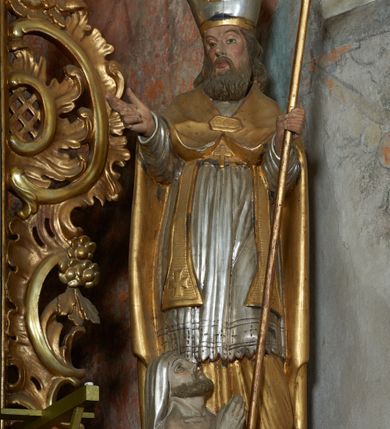 Zdjęcie nr 11: Rzeźba jest drążona i ścięta z tyłu, ustawiona na niskiej podstawie na planie kwadratu. Święty zwrócony jest frontalnie, z głową skierowaną w prawo, w kontrapoście, o odchylonej delikatnie sylwetce. Obie ręce ugięte, rozłożone na boki, w lewej trzyma pastorał, prawą unosi w geście błogosławieństwa.  Twarz ma pociągłą, szczupłą, o migdałowatych oczach, uniesionych brwiach, dużym nosie i pełnych, rozchylonych ustach, okoloną falowaną brodą. Włosy ma brązowe, falowane, sięgające ramion. Na głowie nosi srebrzoną i złoconą infułę z krzyżem kawalerskim w glorii. Święty ubrany jest w złoconą albę, srebrzoną rokietę i złoconą stułę, przepasane w talii. Na piersi ma pektorał, na ramionach zarzuconą złoconą kapę, zapiętą z przodu. Na stopach ma złocone buty. Przy prawej nodze świętego półpostać Piotrowina, zwróconego profilem, z uniesioną głową i dłońmi złożonymi w geście modlitwy. Twarz szczupła, z oczami skierowanymi w górę, okolona krótką brodą. Głowę ma nakrytą srebrzonym całunem opadającym na plecy, tors ma nagi. Polichromia ciała naturalistyczna.
