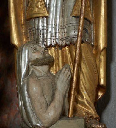 Zdjęcie nr 5: Rzeźba jest drążona i ścięta z tyłu, ustawiona na niskiej podstawie na planie kwadratu. Święty zwrócony jest frontalnie, z głową skierowaną w prawo, w kontrapoście, o odchylonej delikatnie sylwetce. Obie ręce ugięte, rozłożone na boki, w lewej trzyma pastorał, prawą unosi w geście błogosławieństwa.  Twarz ma pociągłą, szczupłą, o migdałowatych oczach, uniesionych brwiach, dużym nosie i pełnych, rozchylonych ustach, okoloną falowaną brodą. Włosy ma brązowe, falowane, sięgające ramion. Na głowie nosi srebrzoną i złoconą infułę z krzyżem kawalerskim w glorii. Święty ubrany jest w złoconą albę, srebrzoną rokietę i złoconą stułę, przepasane w talii. Na piersi ma pektorał, na ramionach zarzuconą złoconą kapę, zapiętą z przodu. Na stopach ma złocone buty. Przy prawej nodze świętego półpostać Piotrowina, zwróconego profilem, z uniesioną głową i dłońmi złożonymi w geście modlitwy. Twarz szczupła, z oczami skierowanymi w górę, okolona krótką brodą. Głowę ma nakrytą srebrzonym całunem opadającym na plecy, tors ma nagi. Polichromia ciała naturalistyczna.
