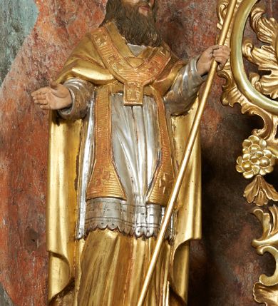 Zdjęcie nr 1: Rzeźba jest drążona i ścięta z tyłu, ustawiona na niskiej podstawie na planie kwadratu. Święty zwrócony jest frontalnie, z głową skierowaną delikatnie w lewo, w kontrapoście. Obie ręce ma ugięte i rozłożone na boki, w lewej trzyma laskę zakończoną krzyżem patriarchalnym. Twarz ma pociągłą, szczupłą, o głęboko osadzonych, migdałowatych oczach, długim nosie i małych ustach, okoloną długą, falowaną brodą, spływającą na pierś. Włosy ma brązowe, falowane, sięgające ramion. Na głowie ma srebrzoną i złoconą infułę z krzyżem. Święty ubrany jest w złoconą albę, srebrzoną rokietę i złoconą stułę, przepasane w talii. Na ramiona ma narzuconą złoconą kapę zapiętą na piesi oraz paliusz. Na stopach ma złocone buty. Polichromia naturalistyczna w partiach ciała.
