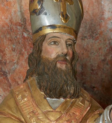 Zdjęcie nr 8: Rzeźba jest drążona i ścięta z tyłu, ustawiona na niskiej podstawie na planie kwadratu. Święty zwrócony jest frontalnie, z głową skierowaną delikatnie w lewo, w kontrapoście. Obie ręce ma ugięte i rozłożone na boki, w lewej trzyma laskę zakończoną krzyżem patriarchalnym. Twarz ma pociągłą, szczupłą, o głęboko osadzonych, migdałowatych oczach, długim nosie i małych ustach, okoloną długą, falowaną brodą, spływającą na pierś. Włosy ma brązowe, falowane, sięgające ramion. Na głowie ma srebrzoną i złoconą infułę z krzyżem. Święty ubrany jest w złoconą albę, srebrzoną rokietę i złoconą stułę, przepasane w talii. Na ramiona ma narzuconą złoconą kapę zapiętą na piesi oraz paliusz. Na stopach ma złocone buty. Polichromia naturalistyczna w partiach ciała.
