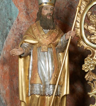 Zdjęcie nr 6: Rzeźba jest drążona i ścięta z tyłu, ustawiona na niskiej podstawie na planie kwadratu. Święty zwrócony jest frontalnie, z głową skierowaną delikatnie w lewo, w kontrapoście. Obie ręce ma ugięte i rozłożone na boki, w lewej trzyma laskę zakończoną krzyżem patriarchalnym. Twarz ma pociągłą, szczupłą, o głęboko osadzonych, migdałowatych oczach, długim nosie i małych ustach, okoloną długą, falowaną brodą, spływającą na pierś. Włosy ma brązowe, falowane, sięgające ramion. Na głowie ma srebrzoną i złoconą infułę z krzyżem. Święty ubrany jest w złoconą albę, srebrzoną rokietę i złoconą stułę, przepasane w talii. Na ramiona ma narzuconą złoconą kapę zapiętą na piesi oraz paliusz. Na stopach ma złocone buty. Polichromia naturalistyczna w partiach ciała.
