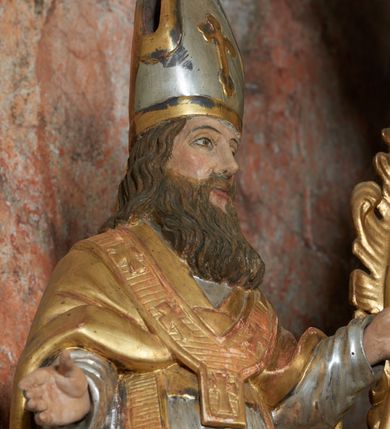 Zdjęcie nr 7: Rzeźba jest drążona i ścięta z tyłu, ustawiona na niskiej podstawie na planie kwadratu. Święty zwrócony jest frontalnie, z głową skierowaną delikatnie w lewo, w kontrapoście. Obie ręce ma ugięte i rozłożone na boki, w lewej trzyma laskę zakończoną krzyżem patriarchalnym. Twarz ma pociągłą, szczupłą, o głęboko osadzonych, migdałowatych oczach, długim nosie i małych ustach, okoloną długą, falowaną brodą, spływającą na pierś. Włosy ma brązowe, falowane, sięgające ramion. Na głowie ma srebrzoną i złoconą infułę z krzyżem. Święty ubrany jest w złoconą albę, srebrzoną rokietę i złoconą stułę, przepasane w talii. Na ramiona ma narzuconą złoconą kapę zapiętą na piesi oraz paliusz. Na stopach ma złocone buty. Polichromia naturalistyczna w partiach ciała.
