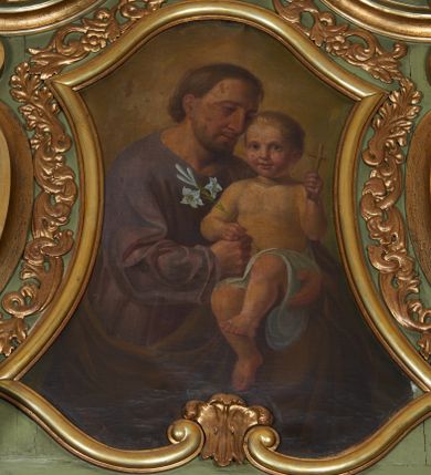 Zdjęcie nr 1: Obraz w formie zbliżonej do trapezu, o wklęsłych bokach, u spodu zamknięty faliście, u góry zaś wypukłą linią. W centrum obrazu przedstawienie św. Józefa w półpostaci, skierowanego w trzech czwartych w lewo, z pochyloną głową. Lewą ręką podtrzymuje Dzieciątko, prawą dłoń, w której trzyma gałązkę lilii kładzie na jego kolanie. Twarz ma pociągłą, o okrągłych, półprzymkniętych oczach, prostym, dużym nosie i pełnych ustach, okoloną jasnobrązową, krótką brodą i włosami. Ubrany jest w fioletową, obszerną tunikę oraz brązowy płaszcz, zarzucony na lewe ramię i otaczający postać. Dzieciątko w pozycji siedzącej, zwrócone w trzech czwartych w prawo, w lewej, uniesionej ręce trzyma krzyżyk, w prawej dłoni położonej na dłoni Józefa gałązkę lilii. Ma okrągłą twarz, o dużych, okrągłych oczach, małym nosie i pełnych ustach, okoloną krótkimi, jasnymi włosami. W partii bioder przepasane jest białą tkaniną. Postacie ukazane na jasnobrązowym tle. 