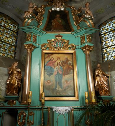 Zdjęcie nr 2: Obraz w formie trapezu o wklęsłych bokach i ściętych dolnych narożach, zamkniętego przerwanym obłokami łukiem odcinkowym nadwieszonym. Święty ukazany w półpostaci, frontalnie, lewą dłoń kładzie na piersi, w prawej opuszczonej wzdłuż ciała trzyma gałązkę lilii. Twarz ma pociągłą, o wąskich oczach, prostym nosie i małych ustach, okoloną krótką siwą brodą i włosami. Wokół głowy nimb. Ubrany jest w luźną zieloną tunikę ze złoconą lamówką oraz przewiązany diagonalnie przez prawe ramię pomarańczowy płaszcz. Postać ukazana na jednolitym czarnym tle. U dołu obrazu złocony napis „«JDŹCIE do JÓZEFA» / (Ks. I Moj. R. 41 W. 55”.
