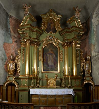 Zdjęcie nr 7: Obraz w formie stojącego prostokąta zamkniętego łukiem odcinkowym. W centrum św. Marcin ukazany w całej postaci, zwrócony w trzech czwartych w prawo. W prawej, wyciągniętej przed siebie i uniesionej ręce podtrzymuje kapę, w lewej, ugiętej pastorał. Twarz ma pociągłą, szczupłą, o starczych rysach, oczach zwróconych w górę, długim nosie oraz niewielkich ustach, okoloną długą, siwą brodą, opadającą na pierś. Włosy ma krótkie, siwe. Na głowie ma malowaną w kolorze srebrnym infułę ze złoconym galonem. Wokół głowy zamknięty nimb. Święty ubrany jest w fioletową sutannę zapinaną na guziki, półprzeźroczystą, białą rokietę, dekorowaną u spodu i przy rękawach koronką, białą, bogato zdobioną w kolorze złotym stułę, fioletowy, zapinany na guziki mucet. Na piersi ma zawieszony pektorał. Na ramiona ma narzuconą malowaną na złoto kapę, z bordową podszewką, zapiętą na piersi. W lewym dolnym rogu obrazu, przed świętym ukazana półpostać mężczyzny, z profilu, z uniesioną głową i wyciągniętą w bok prawą ręką. Twarz ma owalną, z oczami skierowanymi w górę, dużym nosem i otwartymi ustami, z wąsami. Ubrany jest w białą koszulę i ciemne spodnie. Święty stoi na posadzce w szachownicę. Tło jednolite, niebiesko-szare, w górnej części rozświetlone obłoki z wiązkami światła skierowanymi w stronę świętego oraz dwie uskrzydlone główki anielskie. 
