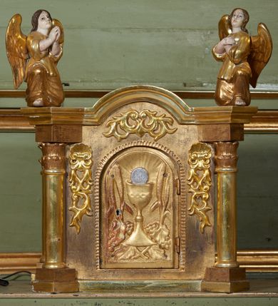 Zdjęcie nr 26: Nastawa ołtarzowa architektoniczna, jednoosiowa, jednokondygnacyjna ze zwieńczeniem, na cokole z czterema prostopadłościennymi postumentami wydzielonymi po bokach, z bramkami. Ołtarz drewniany, prostopadłościenny, z kamienną mensą z portatylem. Antependium drewniane obite miedzianą blachą, w formie leżącego prostokąta, ujętego profilowaną ramą, podzielone na dziesięć pól pasami z ornamentem geometrycznym, oraz aplikami w formie rautów na ich skrzyżowaniu. W środkowym polu monogram Marii oraz dwie apliki z orłem i pogonią u spodu. 
Retabulum flankowane ustawionymi schodkowo i zwróconymi na zewnątrz dwiema parami korynckich kolumn, umieszczonych na tle pilastrów oraz zewnętrzną parą pilastrów dekorowanych kwiatowymi zwisami. Podpory stoją na cokołach, podtrzymują wyłamane belkowanie, przerwane nad polem środkowym, oraz fragmenty przerwanego, falistego przyczółka z siedzącymi na obłokach puttami. W polu środkowym wnęka w formie stojącego prostokąta zamkniętego łukiem półkolistym nadwieszonym z uskokiem w profilowanej ramie, z obrazem św. Marcina. Pole środkowe zamknięte gzymsem przyjmującym kształt łuku odcinkowego nadwieszonego z rokokowym kartuszem z napisem "AVE / MARIA / GRATIA PLENA / LUC. 1.28" oraz z podwieszonymi dwiema różanymi gałązkami. Obraz flankowany dwoma zwisami na wstędze utworzonymi z kwiatów. Kondygnacja ujęta ażurowymi uszami utworzonymi z rocaille'u, kratki i kwiatów. Zwieńczenie w formie zbliżonej po stojącego prostokąta o wklęsłych bokach ujętych spływami wolutowymi z rocaille'm, zamkniętego gzymsem przyjmującym kształt łuku półkolistego nadwieszonego z uskokiem, wyłamanego przy krawędziach, zwieńczonego anthemionem. W polu obraz św. Józefa z Dzieciątkiem w formie zbliżonej do trapezu o wklęsłych bokach i wypukłych podstawach, w ramie. Ujęty z trzech stron wicią roślinną. Bramki na wysokości cokołu nastawy, w formie półkolistych, profilowanych arkad podtrzymywanych na pilastrach o kanelowanych trzonach. Arkady zwieńczone cokołami, na których ustawione są figury św. Wojciecha i św. Stanisława. Struktura polichromowana w kolorze seledynowym, ze złoconymi kolumnami, profilami i ornamentyką.


