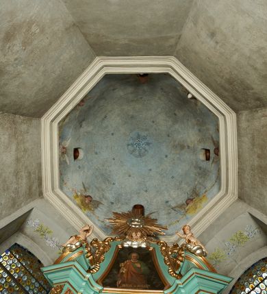 Zdjęcie nr 19: Nastawa ołtarzowa architektoniczna, jednoosiowa, jednokondygnacyjna ze zwieńczeniem, ustawiona na cokole z dwoma prostopadłościennymi postumentami wydzielonymi po bokach, z bramkami w partii cokołu. Ołtarz drewniany, prostopadłościenny, zwężający się ku dołowi, dekorowany z przodu rzeźbionym wieńcem z monogramem Marii, mensa drewniana. Na mensie ustawione tabernakulum w formie drewnianej skrzynki o ściętych przednich narożach, flankowanej parą kolumienek. Drzwiczki w formie stojącego prostokąta dekorowane płyciną. Nastawa ołtarzowa ujęta parą kolumn o stylizowanych kapitelach kompozytowych dźwigających pełne belkowanie, przerwane nad polem środkowym, oraz fragmenty przerwanego, półkolistego przyczółka, na którym klęczą na obłokach dwa putta. Fryz dekorowany ornamentem roślinnym zbliżonym do akantu. W polu głównym wnęka w formie stojącego prostokąta w profilowanej ramie, z obrazem Koronacji Marii. Nad nim medalion utworzony z akantu z monogramem Marii, zwieńczony koroną zamkniętą. Zwieńczenie w formie trapezu, którego brzegi dekorowane są stylizowanym rocaille&#039;m tworzącym uszaki. W polu obraz św. Józefa w formie trapezu o ściętych dolnych narożach, zamkniętego łukiem półkolistym nadwieszonym, w profilowanej ramie, z łukiem częściowo przesłoniętym obłokami z dwoma uskrzydlonymi główkami anielskimi. Nastawa zakończona medalionem z obłoków z Okiem Opatrzności w glorii promienistej. Nastawę flankują bramki w partii cokołu z przejściami w formie stojącego prostokąta zamkniętego łukiem odcinkowym nadwieszonym, zakończone profilowanym gzymsem oraz cokołami dekorowanymi roślinnymi konsolami, na których stoją figury św. Anny i św. Joachima. Struktura polichromowana w kolorze seledynowym, ze złoconymi kolumnami, profilami i  ornamentyką. 
