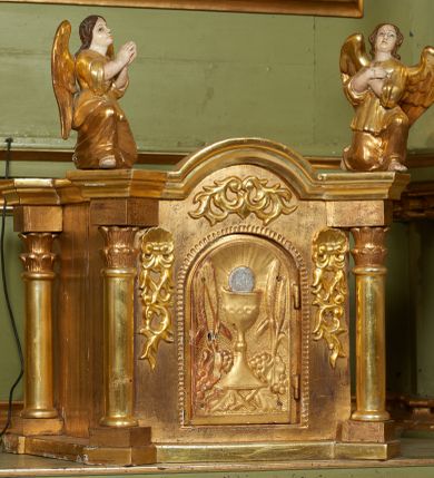 Zdjęcie nr 1: Tabernakulum w formie drewnianej skrzynki ustawionej na niskiej podstawie, ujęte w czterech narożach ukośnie zwróconymi cokołami z kolumnami dźwigającymi imposty oraz profilowany gzyms opasujący całość, nad ścianką frontową przyjmujący kształt łuku odcinkowego. Drzwiczki w formie stojącego prostokąta zamkniętego łukiem półkolistym, dekorowane płaskorzeźbionym przedstawieniem kielicha z hostią, flankowanego winnym gronem i kłosami zbóż. Drzwiczki ujęte z trzech stron ornamentem roślinnym. Na gzymsie dwie figurki klęczących  aniołów. Tabernakulum w całości pozłocone. 

