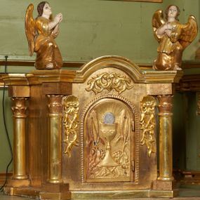 Zdjęcie nr 1: Tabernakulum w formie drewnianej skrzynki ustawionej na niskiej podstawie, ujęte w czterech narożach ukośnie zwróconymi cokołami z kolumnami dźwigającymi imposty oraz profilowany gzyms opasujący całość, nad ścianką frontową przyjmujący kształt łuku odcinkowego. Drzwiczki w formie stojącego prostokąta zamkniętego łukiem półkolistym, dekorowane płaskorzeźbionym przedstawieniem kielicha z hostią, flankowanego winnym gronem i kłosami zbóż. Drzwiczki ujęte z trzech stron ornamentem roślinnym. Na gzymsie dwie figurki klęczących  aniołów. Tabernakulum w całości pozłocone. 

