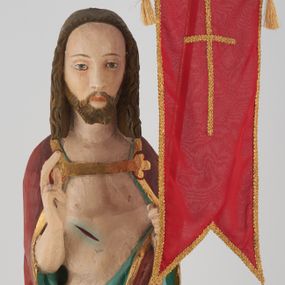 Zdjęcie nr 1: Figura Chrystusa pełnoplastyczna, ustawiona na podstawie na planie ośmioboku oraz na rzeźbionym fragmencie podłoża. Chrystus zwrócony jest frontalnie, w lekkim kontrapoście. Prawą rękę unosi w geście błogosławieństwa, w lewej trzyma drzewce czerwonej chorągwi z tkaniny z naszytym krzyżem, ujętej dwoma chwostami. Twarz ma pociągłą z wysokim czołem, migdałowatymi oczami, wąskim i zadartym nosem oraz małymi ustami, okoloną krótką, falowaną brodą rozdzieloną na dwoje oraz regularnie falowanymi włosami sięgającymi ramion. Ubrany jest w czerwony płaszcz z zieloną podszewką i złoconą lamówką, zapięty na piersi, opadający na plecy i ramiona, zawinięty łukowato z przodu postaci, odsłaniający nagi tors i lewą nogę. Polichromia ciała naturalistyczna z zaznaczonymi śladami męki.