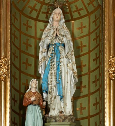 Zdjęcie nr 1: Grupa rzeźbiarska złożona z pełnoplastycznej figury Marii stojącej na cokole i na rzeźbionym fragmencie skały oraz z klęczącej na osobnym cokole i podstawie po prawej stronie Marii pełnoplastycznej figury Bernadety Soubirous. Maria ukazana frontalnie, w całej postaci, w wykroku, z uniesioną głową oraz rękami złożonymi w geście modlitwy. Twarz ma pociągłą, o łagodnych rysach, oczach skierowanych w górę, prostym nosie oraz małych ustach. Ubrana jest w białą, długą suknię, dekorowaną złoconymi rozetami oraz lamówką, przepasaną w talii niebieską szarfą ze złoconymi gwiazdkami i brzegami, zawiązaną z przodu. Na głowę i ramiona ma narzucony biały płaszcz zdobiony złoconymi rozetami i wicią roślinną przy brzegach, ze srebrzonymi pasami na podszewce, spływający kaskadowo po bokach postaci. Na głowie ma złoconą koronę otwartą, wokół niej nimb gwieździsty. Na przedramionach ma zawieszony różaniec. Na stopach złocone róże. Bernadeta zwrócona jest w trzech czwartych w lewo, z uniesioną głową, w lewej dłoni trzyma krzyż z różańca Marii, prawą kładzie na piersi. Twarz ma owalną, o młodzieńczych rysach, migdałowatych oczach, prostym nosie i małych ustach. Ubrana jest w brązową suknię z zieloną zapaską przewiązaną w talii oraz zarzuconą na plecy i skrzyżowaną na piersi jasnobrązową chustę w kratę, na głowie ma biały welon. 