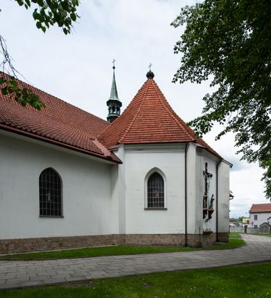 Zdjęcie nr 13: Kościół murowany z cegły i kamienia, tynkowany, orientowany, jednonawowy z kaplicami. Prezbiterium wydłużone, zamknięte trójbocznie, odchylone na południe od osi nawy, przy północnej ścianie niewielka, prostokątna dawna zakrystia (obecnie kruchta), przy południowej dobudowana wtórnie piętrowa dwuczęściowa zakrystia ze skarbcem, od wschodu, na zewnętrznej ścianie kościoła Ogrojec. Nawa główna szersza od prezbiterium, na rzucie prostokąta, od południa wieloboczna kaplica Najświętszego Serca Pana Jezusa (dawniej Przemienienia Pańskiego), od północy i południa dobudowane kaplice boczne na planie prostokąta, północna dwukrotnie większa od południowej, od zachodu wieża na planie kwadratu. Prezbiterium i nawa nakryte sklepieniami pozornymi, oddzielone od ścian gzymsem przebiegającym przez całe wnętrze, łuk tęczowy półkolisty, wewnątrz ujęty pilastrami. Dawne, ostrołukowe okna znajdują się w ścianach nawy głównej. Dostęp do kaplicy i naw bocznych z nawy głównej przez wysokie przejścia arkadowe, w północnej części nawy ujęte opaskami tynkowymi, w północno-zachodniej przejście z barokowym, kamiennym portalem (dawne wejście do kościoła). W starej zakrystii dwa gotyckie, kamienne, ostrołukowe portale oraz gotyckie okno. W wielobocznej kaplicy sklepienie kolebkowe z lunetami, w kaplicach bocznych oraz w nowej zakrystii stropy żelbetowe i sufity tynkowe. Elewacje tynkowane, z obłożoną kamieniem partią cokołu, ściana północna dawnej zakrystii z odsłoniętym wątkiem kamiennym oraz narożną przyporą, wieloboczna kaplica oszkarpowana w narożach. Do wschodniej ściany prezbiterium dobudowany kamienny Ogrójec z arkadą, zwieńczony gzymsem oraz schodkowym szczytem z kamiennymi figurami. Wejścia do kościoła przez kaplice boczne od północy i zachodu oraz przez kruchtę w przyziemiu wieży. Okna w formie stojącego prostokąta w ścianach kaplic bocznych, nowej zakrystii, prezbiterium, okna ostrołukowe w wielobocznej kaplicy. Wieża smukła, trójkondygnacyjna, bez podziałów architektonicznych z oknami na poziomie każdej kondygnacji, zwieńczona hełmem baniastym. Nawa główna i prezbiterium nakryte dachem dwuspadowym, niższe nawy boczne również. W miejscu połączenia nawy i prezbiterium wieżyczka na sygnaturkę. Kaplica przekryta dachem ostrosłupowym, nowa zakrystia i skarbiec dachem trójspadowym, stara zakrystia pulpitowym. 
