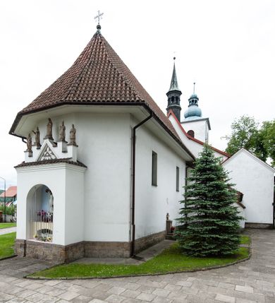 Zdjęcie nr 23: Kościół murowany z cegły i kamienia, tynkowany, orientowany, jednonawowy z kaplicami. Prezbiterium wydłużone, zamknięte trójbocznie, odchylone na południe od osi nawy, przy północnej ścianie niewielka, prostokątna dawna zakrystia (obecnie kruchta), przy południowej dobudowana wtórnie piętrowa dwuczęściowa zakrystia ze skarbcem, od wschodu, na zewnętrznej ścianie kościoła Ogrojec. Nawa główna szersza od prezbiterium, na rzucie prostokąta, od południa wieloboczna kaplica Najświętszego Serca Pana Jezusa (dawniej Przemienienia Pańskiego), od północy i południa dobudowane kaplice boczne na planie prostokąta, północna dwukrotnie większa od południowej, od zachodu wieża na planie kwadratu. Prezbiterium i nawa nakryte sklepieniami pozornymi, oddzielone od ścian gzymsem przebiegającym przez całe wnętrze, łuk tęczowy półkolisty, wewnątrz ujęty pilastrami. Dawne, ostrołukowe okna znajdują się w ścianach nawy głównej. Dostęp do kaplicy i naw bocznych z nawy głównej przez wysokie przejścia arkadowe, w północnej części nawy ujęte opaskami tynkowymi, w północno-zachodniej przejście z barokowym, kamiennym portalem (dawne wejście do kościoła). W starej zakrystii dwa gotyckie, kamienne, ostrołukowe portale oraz gotyckie okno. W wielobocznej kaplicy sklepienie kolebkowe z lunetami, w kaplicach bocznych oraz w nowej zakrystii stropy żelbetowe i sufity tynkowe. Elewacje tynkowane, z obłożoną kamieniem partią cokołu, ściana północna dawnej zakrystii z odsłoniętym wątkiem kamiennym oraz narożną przyporą, wieloboczna kaplica oszkarpowana w narożach. Do wschodniej ściany prezbiterium dobudowany kamienny Ogrójec z arkadą, zwieńczony gzymsem oraz schodkowym szczytem z kamiennymi figurami. Wejścia do kościoła przez kaplice boczne od północy i zachodu oraz przez kruchtę w przyziemiu wieży. Okna w formie stojącego prostokąta w ścianach kaplic bocznych, nowej zakrystii, prezbiterium, okna ostrołukowe w wielobocznej kaplicy. Wieża smukła, trójkondygnacyjna, bez podziałów architektonicznych z oknami na poziomie każdej kondygnacji, zwieńczona hełmem baniastym. Nawa główna i prezbiterium nakryte dachem dwuspadowym, niższe nawy boczne również. W miejscu połączenia nawy i prezbiterium wieżyczka na sygnaturkę. Kaplica przekryta dachem ostrosłupowym, nowa zakrystia i skarbiec dachem trójspadowym, stara zakrystia pulpitowym. 
