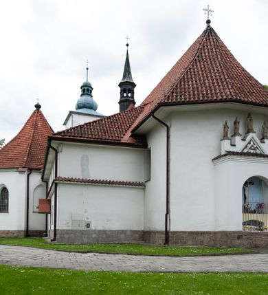 Zdjęcie nr 24: Kościół murowany z cegły i kamienia, tynkowany, orientowany, jednonawowy z kaplicami. Prezbiterium wydłużone, zamknięte trójbocznie, odchylone na południe od osi nawy, przy północnej ścianie niewielka, prostokątna dawna zakrystia (obecnie kruchta), przy południowej dobudowana wtórnie piętrowa dwuczęściowa zakrystia ze skarbcem, od wschodu, na zewnętrznej ścianie kościoła Ogrojec. Nawa główna szersza od prezbiterium, na rzucie prostokąta, od południa wieloboczna kaplica Najświętszego Serca Pana Jezusa (dawniej Przemienienia Pańskiego), od północy i południa dobudowane kaplice boczne na planie prostokąta, północna dwukrotnie większa od południowej, od zachodu wieża na planie kwadratu. Prezbiterium i nawa nakryte sklepieniami pozornymi, oddzielone od ścian gzymsem przebiegającym przez całe wnętrze, łuk tęczowy półkolisty, wewnątrz ujęty pilastrami. Dawne, ostrołukowe okna znajdują się w ścianach nawy głównej. Dostęp do kaplicy i naw bocznych z nawy głównej przez wysokie przejścia arkadowe, w północnej części nawy ujęte opaskami tynkowymi, w północno-zachodniej przejście z barokowym, kamiennym portalem (dawne wejście do kościoła). W starej zakrystii dwa gotyckie, kamienne, ostrołukowe portale oraz gotyckie okno. W wielobocznej kaplicy sklepienie kolebkowe z lunetami, w kaplicach bocznych oraz w nowej zakrystii stropy żelbetowe i sufity tynkowe. Elewacje tynkowane, z obłożoną kamieniem partią cokołu, ściana północna dawnej zakrystii z odsłoniętym wątkiem kamiennym oraz narożną przyporą, wieloboczna kaplica oszkarpowana w narożach. Do wschodniej ściany prezbiterium dobudowany kamienny Ogrójec z arkadą, zwieńczony gzymsem oraz schodkowym szczytem z kamiennymi figurami. Wejścia do kościoła przez kaplice boczne od północy i zachodu oraz przez kruchtę w przyziemiu wieży. Okna w formie stojącego prostokąta w ścianach kaplic bocznych, nowej zakrystii, prezbiterium, okna ostrołukowe w wielobocznej kaplicy. Wieża smukła, trójkondygnacyjna, bez podziałów architektonicznych z oknami na poziomie każdej kondygnacji, zwieńczona hełmem baniastym. Nawa główna i prezbiterium nakryte dachem dwuspadowym, niższe nawy boczne również. W miejscu połączenia nawy i prezbiterium wieżyczka na sygnaturkę. Kaplica przekryta dachem ostrosłupowym, nowa zakrystia i skarbiec dachem trójspadowym, stara zakrystia pulpitowym. 
