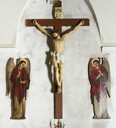 Zdjęcie nr 1: Figura umarłego Chrystusa przybita trzema gwoździami do krzyża o prostym zakończeniu ramion. Ciało w dużym zwisie, o ramionach przyjmujących kształt szerokiego „V”, na lekko ugiętych nogach, ze stopami skrzyżowanymi w układzie prawa na lewą, głowa opada na prawy bark. Twarz ma szczupłą, o dużych, zamkniętych oczach, wąskim nosie i ustach, okoloną krótką brodą i brązowymi włosami, sięgającymi ramion. Na głowie ma koronę cierniową. Ciało ma szczupłe i umięśnione, zwłaszcza w partii ramion i nóg, klatka piersiowa o delikatnie podkreślonej muskulaturze. W prawym boku rana z rzeźbionymi kroplami krwi. Białe, krótkie perizonium zawieszone na podwójnym sznurze, odsłaniające prawe udo, ze zwisem tkaniny przy prawym boku i w partii podbrzusza. Polichromia ciała naturalistyczna z zaznaczonymi śladami męki.  Na zakończeniu pionowej belki krzyża zwój z napisem „I. N. / R. I.”