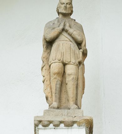 Zdjęcie nr 3: Ogrójec w formie murowanego, tynkowanego wykusza z wnęką w formie stojącego prostokąta zamkniętego łukiem odcinkowym; ustawiony na kamiennej podmurówce, zakończony profilowanym gzymsem i daszkiem okapowym. Ogrójec wieńczy szczyt w formie cokołu z wyodrębnionymi pięcioma postumentami w układzie schodkowym, na których stoją kamienne figury. Pośrodku cokołu rzeźbione w zaprawie Oko Opatrzności w trójkątnym polu, ujętym profilowaną opaską tynkową. Na postumentach od lewej figury św. Ludwika, św. Stanisława z Piotrowinem, Matki Boskiej Niepokalanie Poczętej, św. Wojciecha i św. Walentego. 
Święty Ludwik ukazany frontalnie, z rękami złożonymi w geście modlitwy. Twarz ma okoloną krótkimi włosami i brodą. Ubrany jest w krótką tunikę i spodnie, na nogach ma wysokie buty. Na ramiona ma narzucony płaszcz, opadający na plecy, zaś przez tors przewiązany pas z mieczem, znajdującym się przy lewym boku, na głowie korona zamknięta. 
Święty Stanisław ukazany frontalnie, obie ręce ma ugięte i wyciągnięte do przodu, w lewej trzyma pastorał. Twarz ma okoloną krótkimi włosami. Ubrany jest w albę, rokietę i kapę, na piersi ma krzyż pektoralny, zaś na głowie infułę. U jego nóg leżąca półpostać Piotrowina. 
Matka Boska ukazana frontalnie, stoi na półkuli z wężem, obie dłonie ma złożone na piersi. Twarz ma okoloną długimi włosami, opadającymi na ramiona. Ubrana jest w suknię przepasaną w talii oraz płaszcz zapięty na piersi i opadający kaskadowo po bokach postaci. 
Święty Wojciech ukazany frontalnie, w kontrapoście, w lewej ręce trzyma laskę zakończoną krzyżem, prawą unosi w geście błogosławieństwa. Twarz ma okoloną krótką bodą i włosami. Ubrany jest w albę, rokietę, stułę i kapę, na piersi ma krzyż pektoralny, na dłoniach rękawiczki, a na głowie infułę z krzyżem. 
Święty Walenty ukazany frontalnie, w obu ugiętych rękach trzyma z przodu otwartą księgę. Twarz ma okoloną krótkimi włosami. Ubrany jest w sutannę, komżę, stułę, a na głowie ma biret. 
W dolnej części wnęki Ogrójca nisza w formie leżącego prostokąta zamkniętego łukiem odcinkowym, wewnątrz figura Chrystusa do grobu. Ponad nią zawieszony krucyfiks.
