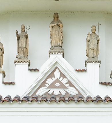 Zdjęcie nr 2: Ogrójec w formie murowanego, tynkowanego wykusza z wnęką w formie stojącego prostokąta zamkniętego łukiem odcinkowym; ustawiony na kamiennej podmurówce, zakończony profilowanym gzymsem i daszkiem okapowym. Ogrójec wieńczy szczyt w formie cokołu z wyodrębnionymi pięcioma postumentami w układzie schodkowym, na których stoją kamienne figury. Pośrodku cokołu rzeźbione w zaprawie Oko Opatrzności w trójkątnym polu, ujętym profilowaną opaską tynkową. Na postumentach od lewej figury św. Ludwika, św. Stanisława z Piotrowinem, Matki Boskiej Niepokalanie Poczętej, św. Wojciecha i św. Walentego. 
Święty Ludwik ukazany frontalnie, z rękami złożonymi w geście modlitwy. Twarz ma okoloną krótkimi włosami i brodą. Ubrany jest w krótką tunikę i spodnie, na nogach ma wysokie buty. Na ramiona ma narzucony płaszcz, opadający na plecy, zaś przez tors przewiązany pas z mieczem, znajdującym się przy lewym boku, na głowie korona zamknięta. 
Święty Stanisław ukazany frontalnie, obie ręce ma ugięte i wyciągnięte do przodu, w lewej trzyma pastorał. Twarz ma okoloną krótkimi włosami. Ubrany jest w albę, rokietę i kapę, na piersi ma krzyż pektoralny, zaś na głowie infułę. U jego nóg leżąca półpostać Piotrowina. 
Matka Boska ukazana frontalnie, stoi na półkuli z wężem, obie dłonie ma złożone na piersi. Twarz ma okoloną długimi włosami, opadającymi na ramiona. Ubrana jest w suknię przepasaną w talii oraz płaszcz zapięty na piersi i opadający kaskadowo po bokach postaci. 
Święty Wojciech ukazany frontalnie, w kontrapoście, w lewej ręce trzyma laskę zakończoną krzyżem, prawą unosi w geście błogosławieństwa. Twarz ma okoloną krótką bodą i włosami. Ubrany jest w albę, rokietę, stułę i kapę, na piersi ma krzyż pektoralny, na dłoniach rękawiczki, a na głowie infułę z krzyżem. 
Święty Walenty ukazany frontalnie, w obu ugiętych rękach trzyma z przodu otwartą księgę. Twarz ma okoloną krótkimi włosami. Ubrany jest w sutannę, komżę, stułę, a na głowie ma biret. 
W dolnej części wnęki Ogrójca nisza w formie leżącego prostokąta zamkniętego łukiem odcinkowym, wewnątrz figura Chrystusa do grobu. Ponad nią zawieszony krucyfiks.
