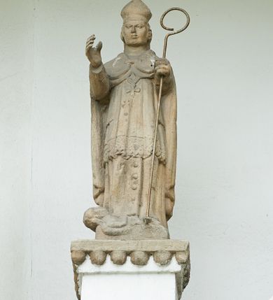 Zdjęcie nr 5: Ogrójec w formie murowanego, tynkowanego wykusza z wnęką w formie stojącego prostokąta zamkniętego łukiem odcinkowym; ustawiony na kamiennej podmurówce, zakończony profilowanym gzymsem i daszkiem okapowym. Ogrójec wieńczy szczyt w formie cokołu z wyodrębnionymi pięcioma postumentami w układzie schodkowym, na których stoją kamienne figury. Pośrodku cokołu rzeźbione w zaprawie Oko Opatrzności w trójkątnym polu, ujętym profilowaną opaską tynkową. Na postumentach od lewej figury św. Ludwika, św. Stanisława z Piotrowinem, Matki Boskiej Niepokalanie Poczętej, św. Wojciecha i św. Walentego. 
Święty Ludwik ukazany frontalnie, z rękami złożonymi w geście modlitwy. Twarz ma okoloną krótkimi włosami i brodą. Ubrany jest w krótką tunikę i spodnie, na nogach ma wysokie buty. Na ramiona ma narzucony płaszcz, opadający na plecy, zaś przez tors przewiązany pas z mieczem, znajdującym się przy lewym boku, na głowie korona zamknięta. 
Święty Stanisław ukazany frontalnie, obie ręce ma ugięte i wyciągnięte do przodu, w lewej trzyma pastorał. Twarz ma okoloną krótkimi włosami. Ubrany jest w albę, rokietę i kapę, na piersi ma krzyż pektoralny, zaś na głowie infułę. U jego nóg leżąca półpostać Piotrowina. 
Matka Boska ukazana frontalnie, stoi na półkuli z wężem, obie dłonie ma złożone na piersi. Twarz ma okoloną długimi włosami, opadającymi na ramiona. Ubrana jest w suknię przepasaną w talii oraz płaszcz zapięty na piersi i opadający kaskadowo po bokach postaci. 
Święty Wojciech ukazany frontalnie, w kontrapoście, w lewej ręce trzyma laskę zakończoną krzyżem, prawą unosi w geście błogosławieństwa. Twarz ma okoloną krótką bodą i włosami. Ubrany jest w albę, rokietę, stułę i kapę, na piersi ma krzyż pektoralny, na dłoniach rękawiczki, a na głowie infułę z krzyżem. 
Święty Walenty ukazany frontalnie, w obu ugiętych rękach trzyma z przodu otwartą księgę. Twarz ma okoloną krótkimi włosami. Ubrany jest w sutannę, komżę, stułę, a na głowie ma biret. 
W dolnej części wnęki Ogrójca nisza w formie leżącego prostokąta zamkniętego łukiem odcinkowym, wewnątrz figura Chrystusa do grobu. Ponad nią zawieszony krucyfiks.
