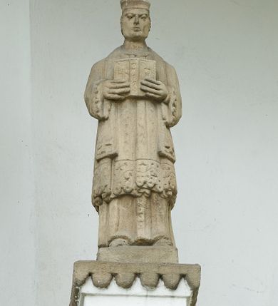 Zdjęcie nr 7: Ogrójec w formie murowanego, tynkowanego wykusza z wnęką w formie stojącego prostokąta zamkniętego łukiem odcinkowym; ustawiony na kamiennej podmurówce, zakończony profilowanym gzymsem i daszkiem okapowym. Ogrójec wieńczy szczyt w formie cokołu z wyodrębnionymi pięcioma postumentami w układzie schodkowym, na których stoją kamienne figury. Pośrodku cokołu rzeźbione w zaprawie Oko Opatrzności w trójkątnym polu, ujętym profilowaną opaską tynkową. Na postumentach od lewej figury św. Ludwika, św. Stanisława z Piotrowinem, Matki Boskiej Niepokalanie Poczętej, św. Wojciecha i św. Walentego. 
Święty Ludwik ukazany frontalnie, z rękami złożonymi w geście modlitwy. Twarz ma okoloną krótkimi włosami i brodą. Ubrany jest w krótką tunikę i spodnie, na nogach ma wysokie buty. Na ramiona ma narzucony płaszcz, opadający na plecy, zaś przez tors przewiązany pas z mieczem, znajdującym się przy lewym boku, na głowie korona zamknięta. 
Święty Stanisław ukazany frontalnie, obie ręce ma ugięte i wyciągnięte do przodu, w lewej trzyma pastorał. Twarz ma okoloną krótkimi włosami. Ubrany jest w albę, rokietę i kapę, na piersi ma krzyż pektoralny, zaś na głowie infułę. U jego nóg leżąca półpostać Piotrowina. 
Matka Boska ukazana frontalnie, stoi na półkuli z wężem, obie dłonie ma złożone na piersi. Twarz ma okoloną długimi włosami, opadającymi na ramiona. Ubrana jest w suknię przepasaną w talii oraz płaszcz zapięty na piersi i opadający kaskadowo po bokach postaci. 
Święty Wojciech ukazany frontalnie, w kontrapoście, w lewej ręce trzyma laskę zakończoną krzyżem, prawą unosi w geście błogosławieństwa. Twarz ma okoloną krótką bodą i włosami. Ubrany jest w albę, rokietę, stułę i kapę, na piersi ma krzyż pektoralny, na dłoniach rękawiczki, a na głowie infułę z krzyżem. 
Święty Walenty ukazany frontalnie, w obu ugiętych rękach trzyma z przodu otwartą księgę. Twarz ma okoloną krótkimi włosami. Ubrany jest w sutannę, komżę, stułę, a na głowie ma biret. 
W dolnej części wnęki Ogrójca nisza w formie leżącego prostokąta zamkniętego łukiem odcinkowym, wewnątrz figura Chrystusa do grobu. Ponad nią zawieszony krucyfiks.
