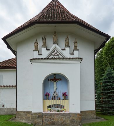 Zdjęcie nr 1: Ogrójec w formie murowanego, tynkowanego wykusza z wnęką w formie stojącego prostokąta zamkniętego łukiem odcinkowym; ustawiony na kamiennej podmurówce, zakończony profilowanym gzymsem i daszkiem okapowym. Ogrójec wieńczy szczyt w formie cokołu z wyodrębnionymi pięcioma postumentami w układzie schodkowym, na których stoją kamienne figury. Pośrodku cokołu rzeźbione w zaprawie Oko Opatrzności w trójkątnym polu, ujętym profilowaną opaską tynkową. Na postumentach od lewej figury św. Ludwika, św. Stanisława z Piotrowinem, Matki Boskiej Niepokalanie Poczętej, św. Wojciecha i św. Walentego. 
Święty Ludwik ukazany frontalnie, z rękami złożonymi w geście modlitwy. Twarz ma okoloną krótkimi włosami i brodą. Ubrany jest w krótką tunikę i spodnie, na nogach ma wysokie buty. Na ramiona ma narzucony płaszcz, opadający na plecy, zaś przez tors przewiązany pas z mieczem, znajdującym się przy lewym boku, na głowie korona zamknięta. 
Święty Stanisław ukazany frontalnie, obie ręce ma ugięte i wyciągnięte do przodu, w lewej trzyma pastorał. Twarz ma okoloną krótkimi włosami. Ubrany jest w albę, rokietę i kapę, na piersi ma krzyż pektoralny, zaś na głowie infułę. U jego nóg leżąca półpostać Piotrowina. 
Matka Boska ukazana frontalnie, stoi na półkuli z wężem, obie dłonie ma złożone na piersi. Twarz ma okoloną długimi włosami, opadającymi na ramiona. Ubrana jest w suknię przepasaną w talii oraz płaszcz zapięty na piersi i opadający kaskadowo po bokach postaci. 
Święty Wojciech ukazany frontalnie, w kontrapoście, w lewej ręce trzyma laskę zakończoną krzyżem, prawą unosi w geście błogosławieństwa. Twarz ma okoloną krótką bodą i włosami. Ubrany jest w albę, rokietę, stułę i kapę, na piersi ma krzyż pektoralny, na dłoniach rękawiczki, a na głowie infułę z krzyżem. 
Święty Walenty ukazany frontalnie, w obu ugiętych rękach trzyma z przodu otwartą księgę. Twarz ma okoloną krótkimi włosami. Ubrany jest w sutannę, komżę, stułę, a na głowie ma biret. 
W dolnej części wnęki Ogrójca nisza w formie leżącego prostokąta zamkniętego łukiem odcinkowym, wewnątrz figura Chrystusa do grobu. Ponad nią zawieszony krucyfiks.
