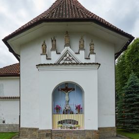 Zdjęcie nr 1: Ogrójec w formie murowanego, tynkowanego wykusza z wnęką w formie stojącego prostokąta zamkniętego łukiem odcinkowym; ustawiony na kamiennej podmurówce, zakończony profilowanym gzymsem i daszkiem okapowym. Ogrójec wieńczy szczyt w formie cokołu z wyodrębnionymi pięcioma postumentami w układzie schodkowym, na których stoją kamienne figury. Pośrodku cokołu rzeźbione w zaprawie Oko Opatrzności w trójkątnym polu, ujętym profilowaną opaską tynkową. Na postumentach od lewej figury św. Ludwika, św. Stanisława z Piotrowinem, Matki Boskiej Niepokalanie Poczętej, św. Wojciecha i św. Walentego. 
Święty Ludwik ukazany frontalnie, z rękami złożonymi w geście modlitwy. Twarz ma okoloną krótkimi włosami i brodą. Ubrany jest w krótką tunikę i spodnie, na nogach ma wysokie buty. Na ramiona ma narzucony płaszcz, opadający na plecy, zaś przez tors przewiązany pas z mieczem, znajdującym się przy lewym boku, na głowie korona zamknięta. 
Święty Stanisław ukazany frontalnie, obie ręce ma ugięte i wyciągnięte do przodu, w lewej trzyma pastorał. Twarz ma okoloną krótkimi włosami. Ubrany jest w albę, rokietę i kapę, na piersi ma krzyż pektoralny, zaś na głowie infułę. U jego nóg leżąca półpostać Piotrowina. 
Matka Boska ukazana frontalnie, stoi na półkuli z wężem, obie dłonie ma złożone na piersi. Twarz ma okoloną długimi włosami, opadającymi na ramiona. Ubrana jest w suknię przepasaną w talii oraz płaszcz zapięty na piersi i opadający kaskadowo po bokach postaci. 
Święty Wojciech ukazany frontalnie, w kontrapoście, w lewej ręce trzyma laskę zakończoną krzyżem, prawą unosi w geście błogosławieństwa. Twarz ma okoloną krótką bodą i włosami. Ubrany jest w albę, rokietę, stułę i kapę, na piersi ma krzyż pektoralny, na dłoniach rękawiczki, a na głowie infułę z krzyżem. 
Święty Walenty ukazany frontalnie, w obu ugiętych rękach trzyma z przodu otwartą księgę. Twarz ma okoloną krótkimi włosami. Ubrany jest w sutannę, komżę, stułę, a na głowie ma biret. 
W dolnej części wnęki Ogrójca nisza w formie leżącego prostokąta zamkniętego łukiem odcinkowym, wewnątrz figura Chrystusa do grobu. Ponad nią zawieszony krucyfiks.
