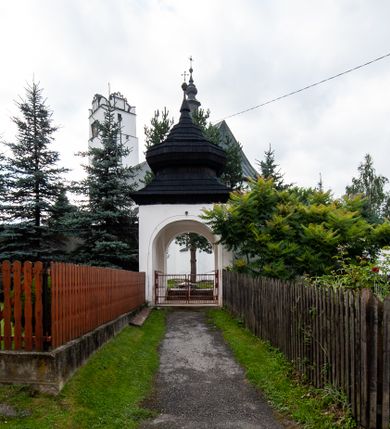 Zdjęcie nr 10: Kościół orientowany, murowany z kamienia, jednonawowy, z prostokątną, trójprzęsłową nawą oraz węższym dwuprzęsłowym prezbiterium zamkniętym trójbocznie, z niewielką prostokątną zakrystią przy północnej ścianie. Od zachodu wieża na planie kwadratu, do północnej ściany nawy dobudowana kaplica Matki Boskiej Karmelitańskiej, do południowej niewielka kruchta. Prezbiterium sklepione żebrowo z jednym rzeźbionym kroksztynem; żebra pokryte późniejszą dekoracją stiukową. Nawa wtórnie sklepiona kolebką z lunetami na gurtach, rozczłonkowana opilastrowanymi półfilarami. Pilastry i zacheuszki ozdobione stiukową ornamentacją. Do zakrystii prowadzi kamienny portal. Kościół opięty grubymi szkarpami jednouskokowymi, nakrytymi daszkami. W dwóch oknach prezbiterium i jednym nawy od południa kamienne maswerki; w zakrystii od wschodu wąskie okienko, tzw. romańskie. W pólnocno-wschodnim narożniku nawy wtórnie wkomponowany rzeźbiony, bardzo zniszczony kroksztyn. Z południowej kruchty do nawy prowadzi kamienny portal o trzech parach kolumienek dźwigających archiwolty. Nawa i prezbiterium nakryte dachem, na ich styku wieżyczka na sygnaturkę. Wieża smukła, bez podziałów architektonicznych, ściany najwyższej kondygnacji cofnięte w głąb, przy krawędzi kamienne kroksztyny, pozostałe po niezachowanej drewnianej hurdycji. Wieża zwieńczona ceglaną attyką, zasłaniającą dwuspadowy dach. W górnej części jej trzonu okna usytuowane z czterech stron. W miejscu hurdycji, na zachodniej ścianie zegar. 

Kościół otoczony niskim murem kamiennym; od zachodu dzwonnica bramna na planie kwadratu z niesklepioną sienią przejściową, nakryta blaszanym hełmem; od południa i od wschodu bramki nakryte hełmami, przy czym jeden pokryty jest gontem, drugi blachą.