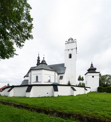 Zdjęcie nr 46: Kościół orientowany, murowany z kamienia, jednonawowy, z prostokątną, trójprzęsłową nawą oraz węższym dwuprzęsłowym prezbiterium zamkniętym trójbocznie, z niewielką prostokątną zakrystią przy północnej ścianie. Od zachodu wieża na planie kwadratu, do północnej ściany nawy dobudowana kaplica Matki Boskiej Karmelitańskiej, do południowej niewielka kruchta. Prezbiterium sklepione żebrowo z jednym rzeźbionym kroksztynem; żebra pokryte późniejszą dekoracją stiukową. Nawa wtórnie sklepiona kolebką z lunetami na gurtach, rozczłonkowana opilastrowanymi półfilarami. Pilastry i zacheuszki ozdobione stiukową ornamentacją. Do zakrystii prowadzi kamienny portal. Kościół opięty grubymi szkarpami jednouskokowymi, nakrytymi daszkami. W dwóch oknach prezbiterium i jednym nawy od południa kamienne maswerki; w zakrystii od wschodu wąskie okienko, tzw. romańskie. W pólnocno-wschodnim narożniku nawy wtórnie wkomponowany rzeźbiony, bardzo zniszczony kroksztyn. Z południowej kruchty do nawy prowadzi kamienny portal o trzech parach kolumienek dźwigających archiwolty. Nawa i prezbiterium nakryte dachem, na ich styku wieżyczka na sygnaturkę. Wieża smukła, bez podziałów architektonicznych, ściany najwyższej kondygnacji cofnięte w głąb, przy krawędzi kamienne kroksztyny, pozostałe po niezachowanej drewnianej hurdycji. Wieża zwieńczona ceglaną attyką, zasłaniającą dwuspadowy dach. W górnej części jej trzonu okna usytuowane z czterech stron. W miejscu hurdycji, na zachodniej ścianie zegar. 

Kościół otoczony niskim murem kamiennym; od zachodu dzwonnica bramna na planie kwadratu z niesklepioną sienią przejściową, nakryta blaszanym hełmem; od południa i od wschodu bramki nakryte hełmami, przy czym jeden pokryty jest gontem, drugi blachą.