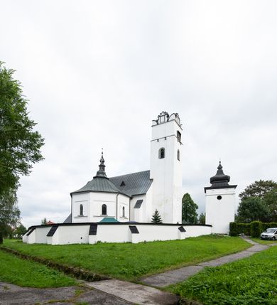 Zdjęcie nr 13: Kościół orientowany, murowany z kamienia, jednonawowy, z prostokątną, trójprzęsłową nawą oraz węższym dwuprzęsłowym prezbiterium zamkniętym trójbocznie, z niewielką prostokątną zakrystią przy północnej ścianie. Od zachodu wieża na planie kwadratu, do północnej ściany nawy dobudowana kaplica Matki Boskiej Karmelitańskiej, do południowej niewielka kruchta. Prezbiterium sklepione żebrowo z jednym rzeźbionym kroksztynem; żebra pokryte późniejszą dekoracją stiukową. Nawa wtórnie sklepiona kolebką z lunetami na gurtach, rozczłonkowana opilastrowanymi półfilarami. Pilastry i zacheuszki ozdobione stiukową ornamentacją. Do zakrystii prowadzi kamienny portal. Kościół opięty grubymi szkarpami jednouskokowymi, nakrytymi daszkami. W dwóch oknach prezbiterium i jednym nawy od południa kamienne maswerki; w zakrystii od wschodu wąskie okienko, tzw. romańskie. W pólnocno-wschodnim narożniku nawy wtórnie wkomponowany rzeźbiony, bardzo zniszczony kroksztyn. Z południowej kruchty do nawy prowadzi kamienny portal o trzech parach kolumienek dźwigających archiwolty. Nawa i prezbiterium nakryte dachem, na ich styku wieżyczka na sygnaturkę. Wieża smukła, bez podziałów architektonicznych, ściany najwyższej kondygnacji cofnięte w głąb, przy krawędzi kamienne kroksztyny, pozostałe po niezachowanej drewnianej hurdycji. Wieża zwieńczona ceglaną attyką, zasłaniającą dwuspadowy dach. W górnej części jej trzonu okna usytuowane z czterech stron. W miejscu hurdycji, na zachodniej ścianie zegar. 

Kościół otoczony niskim murem kamiennym; od zachodu dzwonnica bramna na planie kwadratu z niesklepioną sienią przejściową, nakryta blaszanym hełmem; od południa i od wschodu bramki nakryte hełmami, przy czym jeden pokryty jest gontem, drugi blachą.