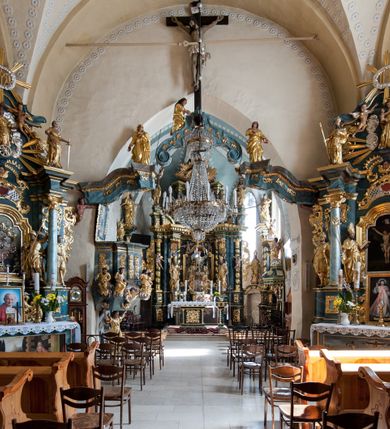 Zdjęcie nr 22: Kościół orientowany, murowany z kamienia, jednonawowy, z prostokątną, trójprzęsłową nawą oraz węższym dwuprzęsłowym prezbiterium zamkniętym trójbocznie, z niewielką prostokątną zakrystią przy północnej ścianie. Od zachodu wieża na planie kwadratu, do północnej ściany nawy dobudowana kaplica Matki Boskiej Karmelitańskiej, do południowej niewielka kruchta. Prezbiterium sklepione żebrowo z jednym rzeźbionym kroksztynem; żebra pokryte późniejszą dekoracją stiukową. Nawa wtórnie sklepiona kolebką z lunetami na gurtach, rozczłonkowana opilastrowanymi półfilarami. Pilastry i zacheuszki ozdobione stiukową ornamentacją. Do zakrystii prowadzi kamienny portal. Kościół opięty grubymi szkarpami jednouskokowymi, nakrytymi daszkami. W dwóch oknach prezbiterium i jednym nawy od południa kamienne maswerki; w zakrystii od wschodu wąskie okienko, tzw. romańskie. W pólnocno-wschodnim narożniku nawy wtórnie wkomponowany rzeźbiony, bardzo zniszczony kroksztyn. Z południowej kruchty do nawy prowadzi kamienny portal o trzech parach kolumienek dźwigających archiwolty. Nawa i prezbiterium nakryte dachem, na ich styku wieżyczka na sygnaturkę. Wieża smukła, bez podziałów architektonicznych, ściany najwyższej kondygnacji cofnięte w głąb, przy krawędzi kamienne kroksztyny, pozostałe po niezachowanej drewnianej hurdycji. Wieża zwieńczona ceglaną attyką, zasłaniającą dwuspadowy dach. W górnej części jej trzonu okna usytuowane z czterech stron. W miejscu hurdycji, na zachodniej ścianie zegar. 

Kościół otoczony niskim murem kamiennym; od zachodu dzwonnica bramna na planie kwadratu z niesklepioną sienią przejściową, nakryta blaszanym hełmem; od południa i od wschodu bramki nakryte hełmami, przy czym jeden pokryty jest gontem, drugi blachą.