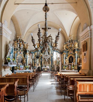 Zdjęcie nr 21: Kościół orientowany, murowany z kamienia, jednonawowy, z prostokątną, trójprzęsłową nawą oraz węższym dwuprzęsłowym prezbiterium zamkniętym trójbocznie, z niewielką prostokątną zakrystią przy północnej ścianie. Od zachodu wieża na planie kwadratu, do północnej ściany nawy dobudowana kaplica Matki Boskiej Karmelitańskiej, do południowej niewielka kruchta. Prezbiterium sklepione żebrowo z jednym rzeźbionym kroksztynem; żebra pokryte późniejszą dekoracją stiukową. Nawa wtórnie sklepiona kolebką z lunetami na gurtach, rozczłonkowana opilastrowanymi półfilarami. Pilastry i zacheuszki ozdobione stiukową ornamentacją. Do zakrystii prowadzi kamienny portal. Kościół opięty grubymi szkarpami jednouskokowymi, nakrytymi daszkami. W dwóch oknach prezbiterium i jednym nawy od południa kamienne maswerki; w zakrystii od wschodu wąskie okienko, tzw. romańskie. W pólnocno-wschodnim narożniku nawy wtórnie wkomponowany rzeźbiony, bardzo zniszczony kroksztyn. Z południowej kruchty do nawy prowadzi kamienny portal o trzech parach kolumienek dźwigających archiwolty. Nawa i prezbiterium nakryte dachem, na ich styku wieżyczka na sygnaturkę. Wieża smukła, bez podziałów architektonicznych, ściany najwyższej kondygnacji cofnięte w głąb, przy krawędzi kamienne kroksztyny, pozostałe po niezachowanej drewnianej hurdycji. Wieża zwieńczona ceglaną attyką, zasłaniającą dwuspadowy dach. W górnej części jej trzonu okna usytuowane z czterech stron. W miejscu hurdycji, na zachodniej ścianie zegar. 

Kościół otoczony niskim murem kamiennym; od zachodu dzwonnica bramna na planie kwadratu z niesklepioną sienią przejściową, nakryta blaszanym hełmem; od południa i od wschodu bramki nakryte hełmami, przy czym jeden pokryty jest gontem, drugi blachą.