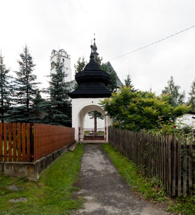 Zdjęcie nr 11: Kościół orientowany, murowany z kamienia, jednonawowy, z prostokątną, trójprzęsłową nawą oraz węższym dwuprzęsłowym prezbiterium zamkniętym trójbocznie, z niewielką prostokątną zakrystią przy północnej ścianie. Od zachodu wieża na planie kwadratu, do północnej ściany nawy dobudowana kaplica Matki Boskiej Karmelitańskiej, do południowej niewielka kruchta. Prezbiterium sklepione żebrowo z jednym rzeźbionym kroksztynem; żebra pokryte późniejszą dekoracją stiukową. Nawa wtórnie sklepiona kolebką z lunetami na gurtach, rozczłonkowana opilastrowanymi półfilarami. Pilastry i zacheuszki ozdobione stiukową ornamentacją. Do zakrystii prowadzi kamienny portal. Kościół opięty grubymi szkarpami jednouskokowymi, nakrytymi daszkami. W dwóch oknach prezbiterium i jednym nawy od południa kamienne maswerki; w zakrystii od wschodu wąskie okienko, tzw. romańskie. W pólnocno-wschodnim narożniku nawy wtórnie wkomponowany rzeźbiony, bardzo zniszczony kroksztyn. Z południowej kruchty do nawy prowadzi kamienny portal o trzech parach kolumienek dźwigających archiwolty. Nawa i prezbiterium nakryte dachem, na ich styku wieżyczka na sygnaturkę. Wieża smukła, bez podziałów architektonicznych, ściany najwyższej kondygnacji cofnięte w głąb, przy krawędzi kamienne kroksztyny, pozostałe po niezachowanej drewnianej hurdycji. Wieża zwieńczona ceglaną attyką, zasłaniającą dwuspadowy dach. W górnej części jej trzonu okna usytuowane z czterech stron. W miejscu hurdycji, na zachodniej ścianie zegar. 

Kościół otoczony niskim murem kamiennym; od zachodu dzwonnica bramna na planie kwadratu z niesklepioną sienią przejściową, nakryta blaszanym hełmem; od południa i od wschodu bramki nakryte hełmami, przy czym jeden pokryty jest gontem, drugi blachą.