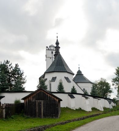 Zdjęcie nr 48: Kościół orientowany, murowany z kamienia, jednonawowy, z prostokątną, trójprzęsłową nawą oraz węższym dwuprzęsłowym prezbiterium zamkniętym trójbocznie, z niewielką prostokątną zakrystią przy północnej ścianie. Od zachodu wieża na planie kwadratu, do północnej ściany nawy dobudowana kaplica Matki Boskiej Karmelitańskiej, do południowej niewielka kruchta. Prezbiterium sklepione żebrowo z jednym rzeźbionym kroksztynem; żebra pokryte późniejszą dekoracją stiukową. Nawa wtórnie sklepiona kolebką z lunetami na gurtach, rozczłonkowana opilastrowanymi półfilarami. Pilastry i zacheuszki ozdobione stiukową ornamentacją. Do zakrystii prowadzi kamienny portal. Kościół opięty grubymi szkarpami jednouskokowymi, nakrytymi daszkami. W dwóch oknach prezbiterium i jednym nawy od południa kamienne maswerki; w zakrystii od wschodu wąskie okienko, tzw. romańskie. W pólnocno-wschodnim narożniku nawy wtórnie wkomponowany rzeźbiony, bardzo zniszczony kroksztyn. Z południowej kruchty do nawy prowadzi kamienny portal o trzech parach kolumienek dźwigających archiwolty. Nawa i prezbiterium nakryte dachem, na ich styku wieżyczka na sygnaturkę. Wieża smukła, bez podziałów architektonicznych, ściany najwyższej kondygnacji cofnięte w głąb, przy krawędzi kamienne kroksztyny, pozostałe po niezachowanej drewnianej hurdycji. Wieża zwieńczona ceglaną attyką, zasłaniającą dwuspadowy dach. W górnej części jej trzonu okna usytuowane z czterech stron. W miejscu hurdycji, na zachodniej ścianie zegar. 

Kościół otoczony niskim murem kamiennym; od zachodu dzwonnica bramna na planie kwadratu z niesklepioną sienią przejściową, nakryta blaszanym hełmem; od południa i od wschodu bramki nakryte hełmami, przy czym jeden pokryty jest gontem, drugi blachą.