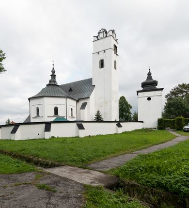 Zdjęcie nr 24: Kościół orientowany, murowany z kamienia, jednonawowy, z prostokątną, trójprzęsłową nawą oraz węższym dwuprzęsłowym prezbiterium zamkniętym trójbocznie, z niewielką prostokątną zakrystią przy północnej ścianie. Od zachodu wieża na planie kwadratu, do północnej ściany nawy dobudowana kaplica Matki Boskiej Karmelitańskiej, do południowej niewielka kruchta. Prezbiterium sklepione żebrowo z jednym rzeźbionym kroksztynem; żebra pokryte późniejszą dekoracją stiukową. Nawa wtórnie sklepiona kolebką z lunetami na gurtach, rozczłonkowana opilastrowanymi półfilarami. Pilastry i zacheuszki ozdobione stiukową ornamentacją. Do zakrystii prowadzi kamienny portal. Kościół opięty grubymi szkarpami jednouskokowymi, nakrytymi daszkami. W dwóch oknach prezbiterium i jednym nawy od południa kamienne maswerki; w zakrystii od wschodu wąskie okienko, tzw. romańskie. W pólnocno-wschodnim narożniku nawy wtórnie wkomponowany rzeźbiony, bardzo zniszczony kroksztyn. Z południowej kruchty do nawy prowadzi kamienny portal o trzech parach kolumienek dźwigających archiwolty. Nawa i prezbiterium nakryte dachem, na ich styku wieżyczka na sygnaturkę. Wieża smukła, bez podziałów architektonicznych, ściany najwyższej kondygnacji cofnięte w głąb, przy krawędzi kamienne kroksztyny, pozostałe po niezachowanej drewnianej hurdycji. Wieża zwieńczona ceglaną attyką, zasłaniającą dwuspadowy dach. W górnej części jej trzonu okna usytuowane z czterech stron. W miejscu hurdycji, na zachodniej ścianie zegar. 

Kościół otoczony niskim murem kamiennym; od zachodu dzwonnica bramna na planie kwadratu z niesklepioną sienią przejściową, nakryta blaszanym hełmem; od południa i od wschodu bramki nakryte hełmami, przy czym jeden pokryty jest gontem, drugi blachą.